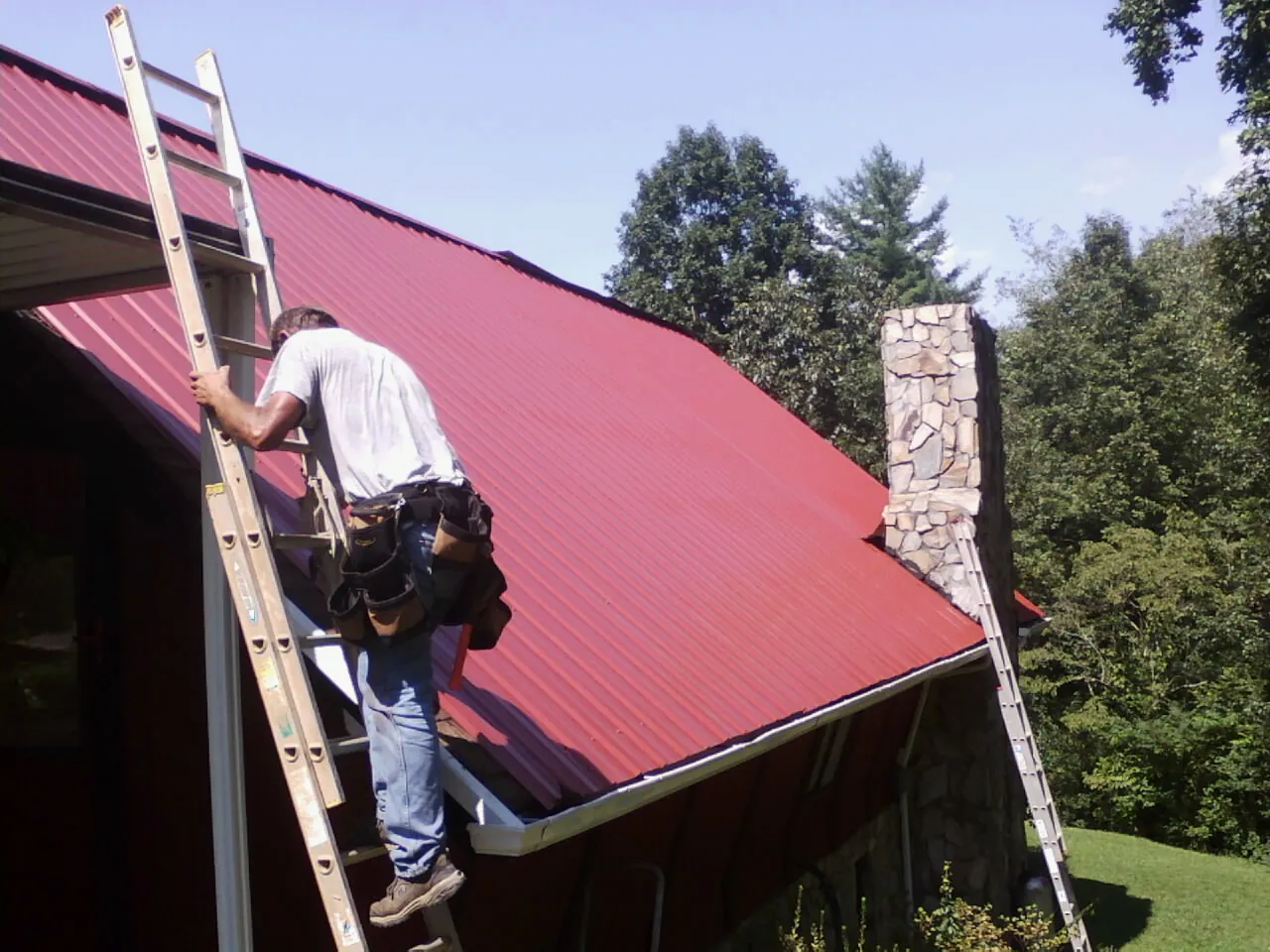 Roofer on a ladder beside a red metal roof and stone chimney — Soffit & Fascia Repair by Roofing Brothers in Pennsauken